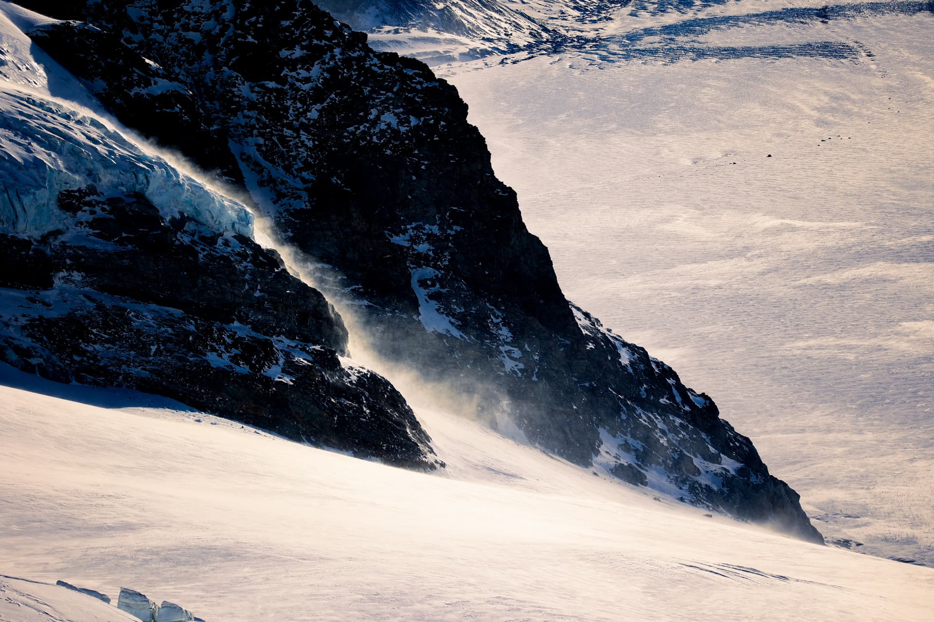 Wind wirbelt Schnee am Fuss einer dunklen Felswand auf